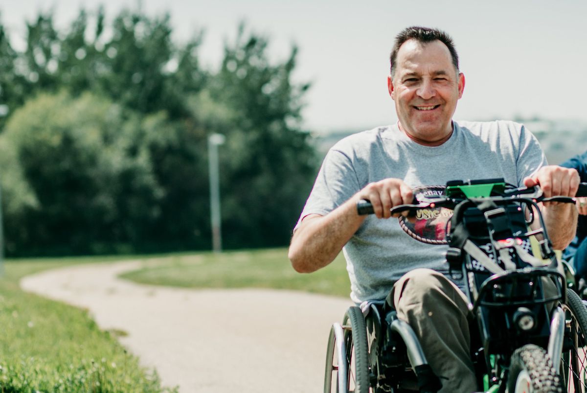 pat-harris-bike SCI BC peer and donor Pat Harris rides a three wheel power wheelchair on a paved pathway in the summer