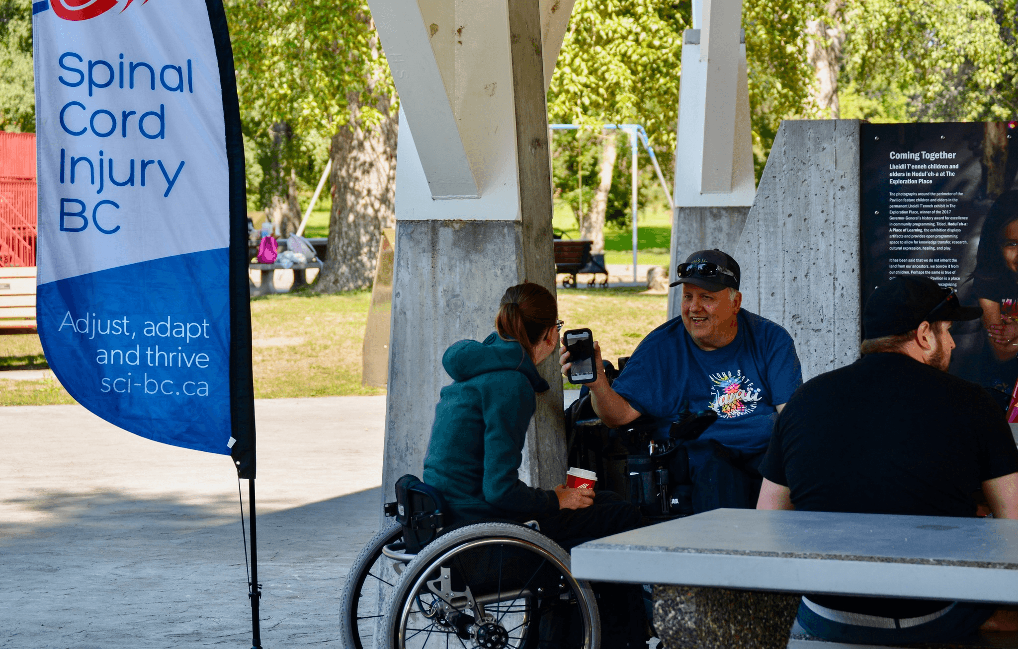 A man and women sitting in wheelchairs beside a picnic table, with a Spinal Cord Injury BC banner to their left. The man is smiling as he shows the woman a text message on his phone screen.